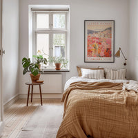 a cozy bedroom with a large bed, a wooden nightstand, and a large window with a plant on the sill. The room has a warm, inviting atmosphere with a neutral color palette and natural elements like the potted plant and the framed poster on the wall.