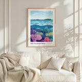 a framed poster of a vibrant underwater scene, with a lush coral reef and a serene ocean backdrop. The poster is hung on a white wall above a beige couch, which is adorned with a throw blanket and a single pillow.