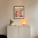 a white cabinet with a potted plant and a lamp on top, set against a wall with a framed poster of a cityscape above it.