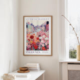 a framed poster of a cityscape with a flower market scene, hanging on a wall in a room with a window and a small table with books and a bowl.