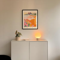 a framed poster of a tropical beach scene hanging on a wall above a white cabinet with a plant and a lamp on top.