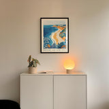 a framed poster of Bondi Beach hanging on a wall above a white cabinet with a plant and a lamp on top.
