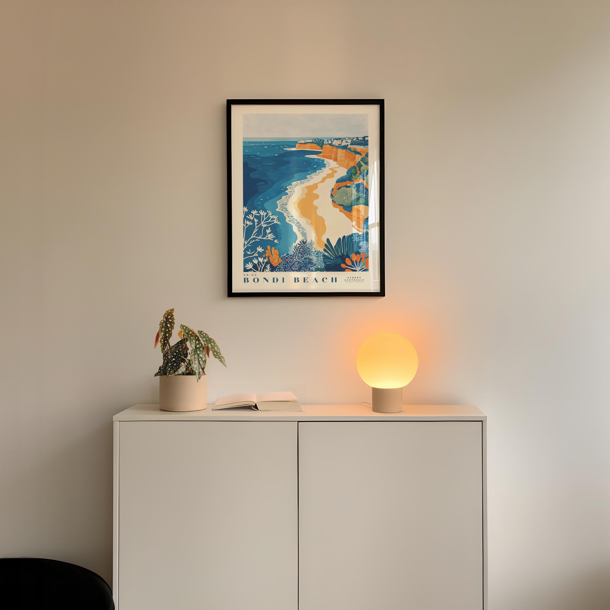 a framed poster of Bondi Beach hanging on a wall above a white cabinet with a plant and a lamp on top.