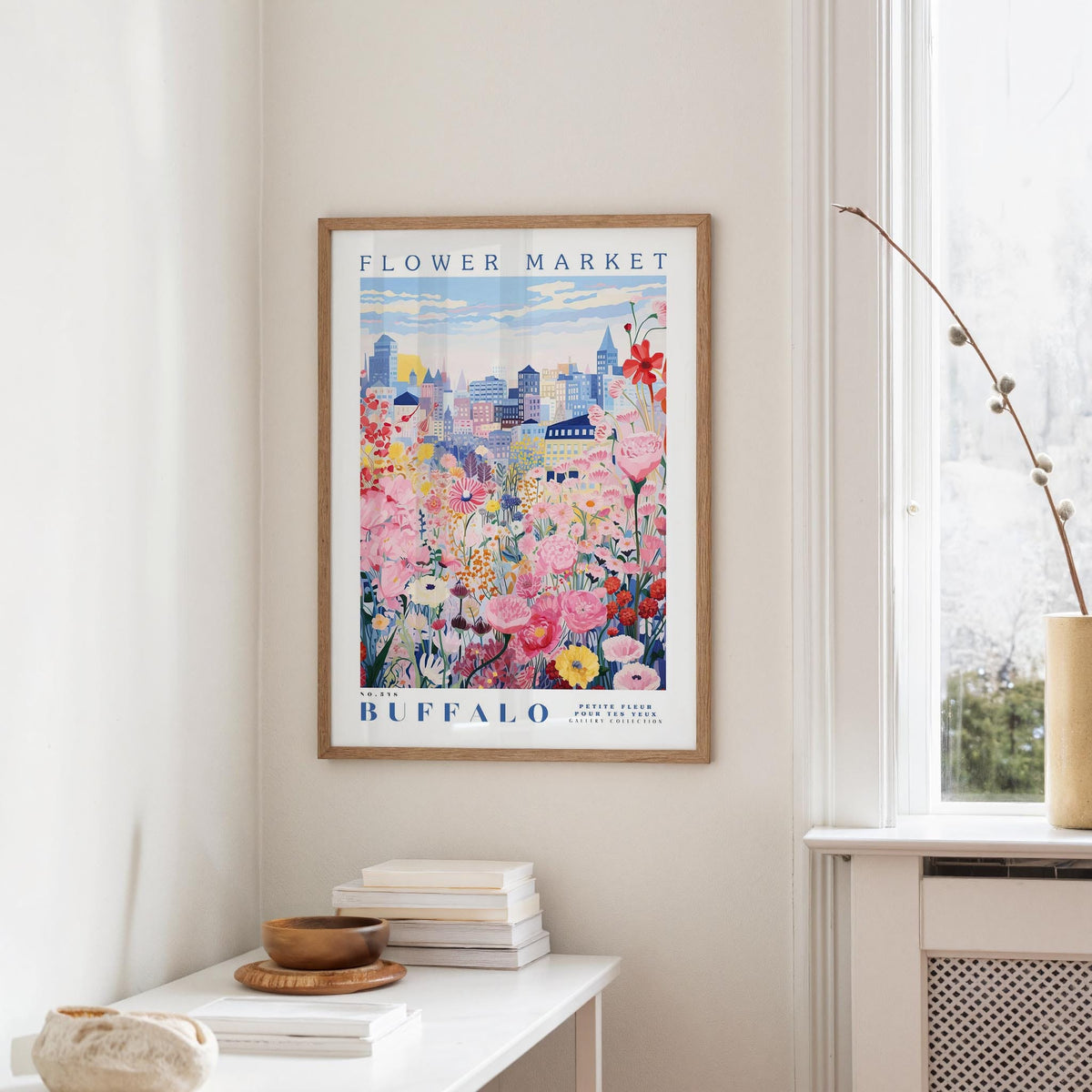 a framed poster of a colorful floral scene hanging on a wall, with a window and a small table with books and a vase nearby.