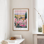 a framed poster of a colorful garden scene with pink flowers, hanging on a wall in a room with a white table and a window.