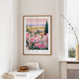 a framed poster of a colorful garden scene with pink flowers, hanging on a wall in a room with a white table and a window.