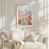 a framed poster of a colorful cityscape with flowers, hanging on a wall above a white couch with pillows.