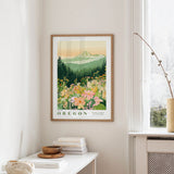 a framed poster of a mountain landscape with a field of flowers, hanging on a wall in a room with a white table and a vase of flowers.