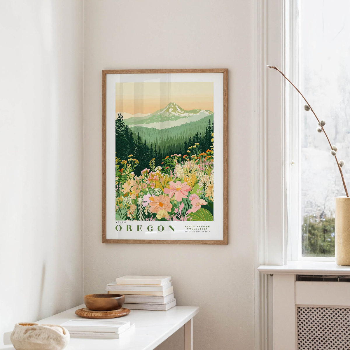 a framed poster of a mountain landscape with a field of flowers, hanging on a wall in a room with a white table and a vase of flowers.