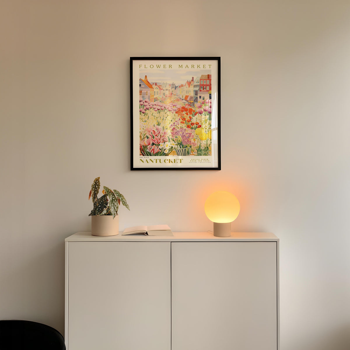 a white cabinet with a potted plant and a lamp on top, set against a wall with a framed poster of a flower market above it.