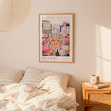 a framed poster of a colorful cityscape with flowers hanging on a wall above a bed with striped pillows.