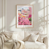 a framed poster of a colorful floral scene hanging on a wall above a white couch with a blanket draped over it.