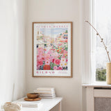a framed poster of a colorful floral scene hanging on a wall, with a white table and a vase of flowers in the foreground.
