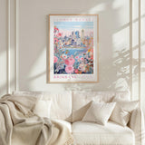 a framed poster of a cityscape with a flower market in the background, hanging on a wall above a white couch with a blanket draped over it.