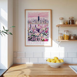 a framed poster of a city skyline with a field of flowers in the foreground, hanging on a wall above a kitchen counter with a bowl of lemons and other kitchen items.