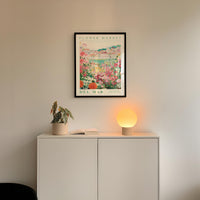 a white cabinet with a potted plant and a lamp on top, set against a wall with a framed poster of a flower market.