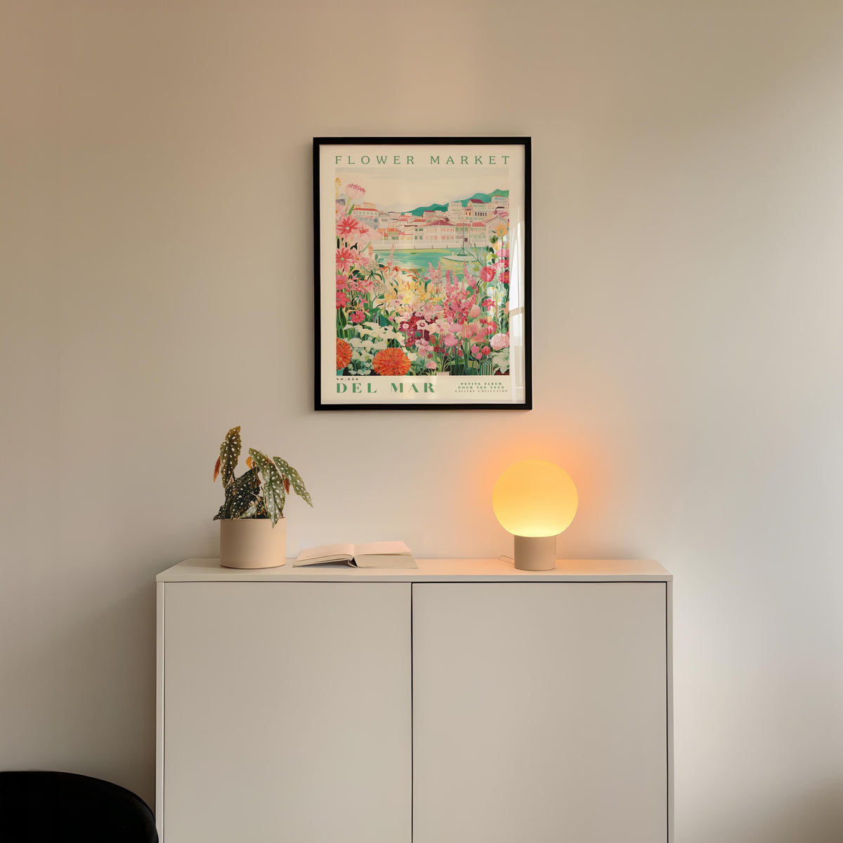 a white cabinet with a potted plant and a lamp on top, set against a wall with a framed poster of a flower market.