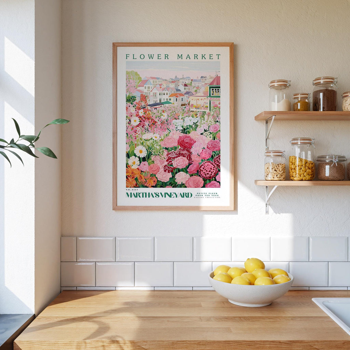 a kitchen scene with a framed poster of a flower market hanging on the wall above a wooden counter. On the counter, there is a bowl of lemons and a plant. The kitchen has white tile walls and a wooden floor.