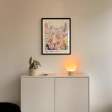 a white cabinet with a potted plant and a lamp on top, set against a wall with a framed poster of a cityscape above it.