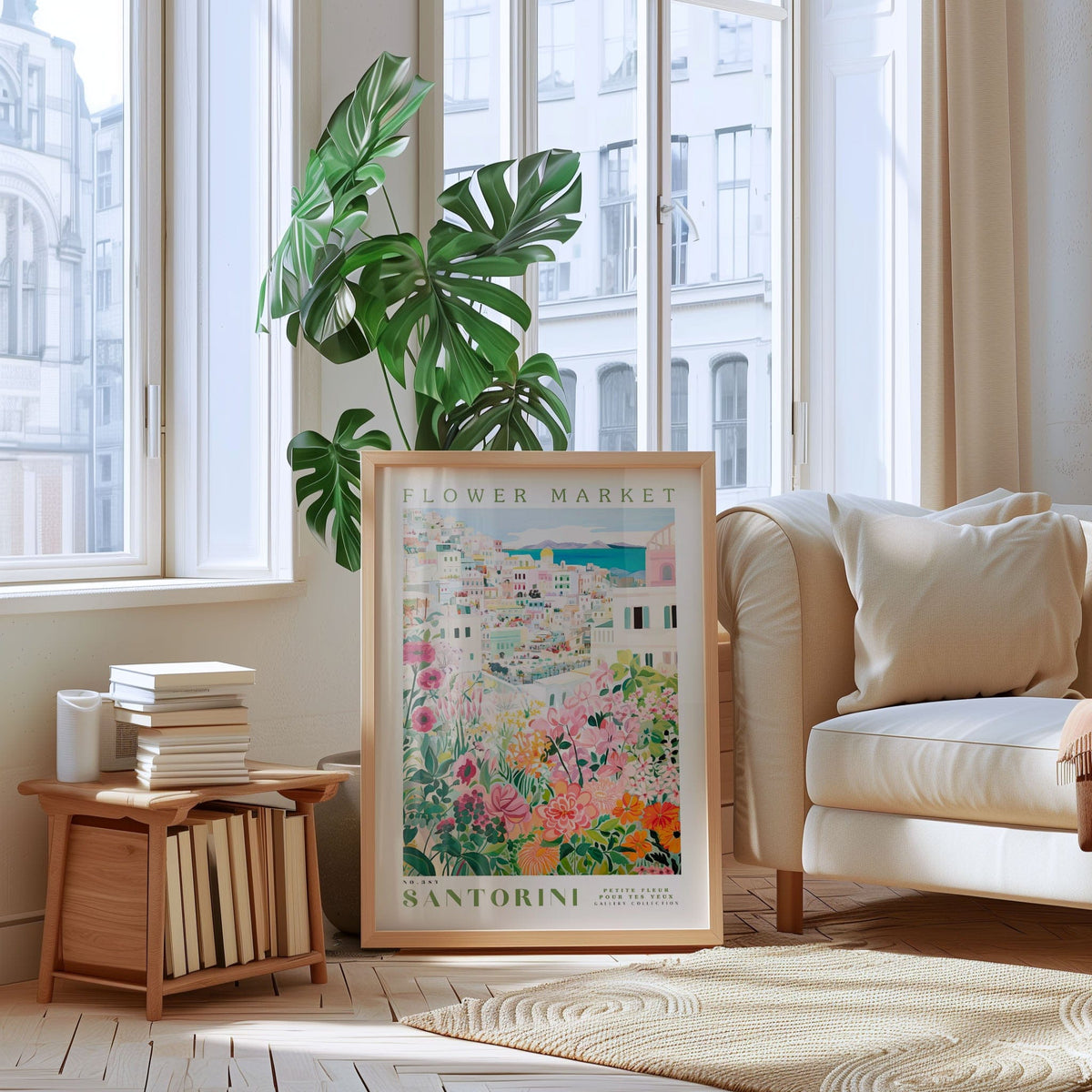 a living room with a large window, a framed poster of Santorini, a stack of books, a white sofa, and a potted plant.