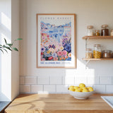 a kitchen scene with a framed poster of a flower market hanging on the wall above a wooden counter. On the counter, there is a bowl of lemons and a plant. The kitchen has white tile backsplash and shelves with various jars and containers.