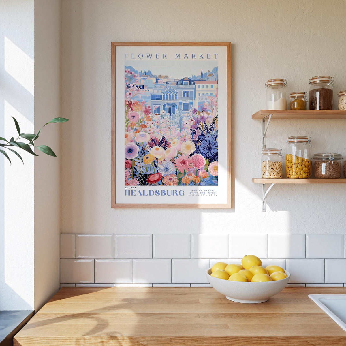a kitchen scene with a framed poster of a flower market hanging on the wall above a wooden counter. On the counter, there is a bowl of lemons and a plant. The kitchen has white tile backsplash and shelves with various jars and containers.