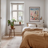 A cozy bedroom with a large bed, a nightstand with a plant, and a framed poster on the wall. The room has a warm, inviting atmosphere with natural light coming through a window.
