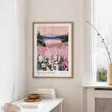 a framed poster of a scenic landscape with a lake and mountains, hanging on a wall in a room with a white table and a vase of flowers.