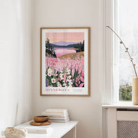 a framed poster of a scenic landscape with a lake and mountains, hanging on a wall in a room with a white table and a vase of flowers.