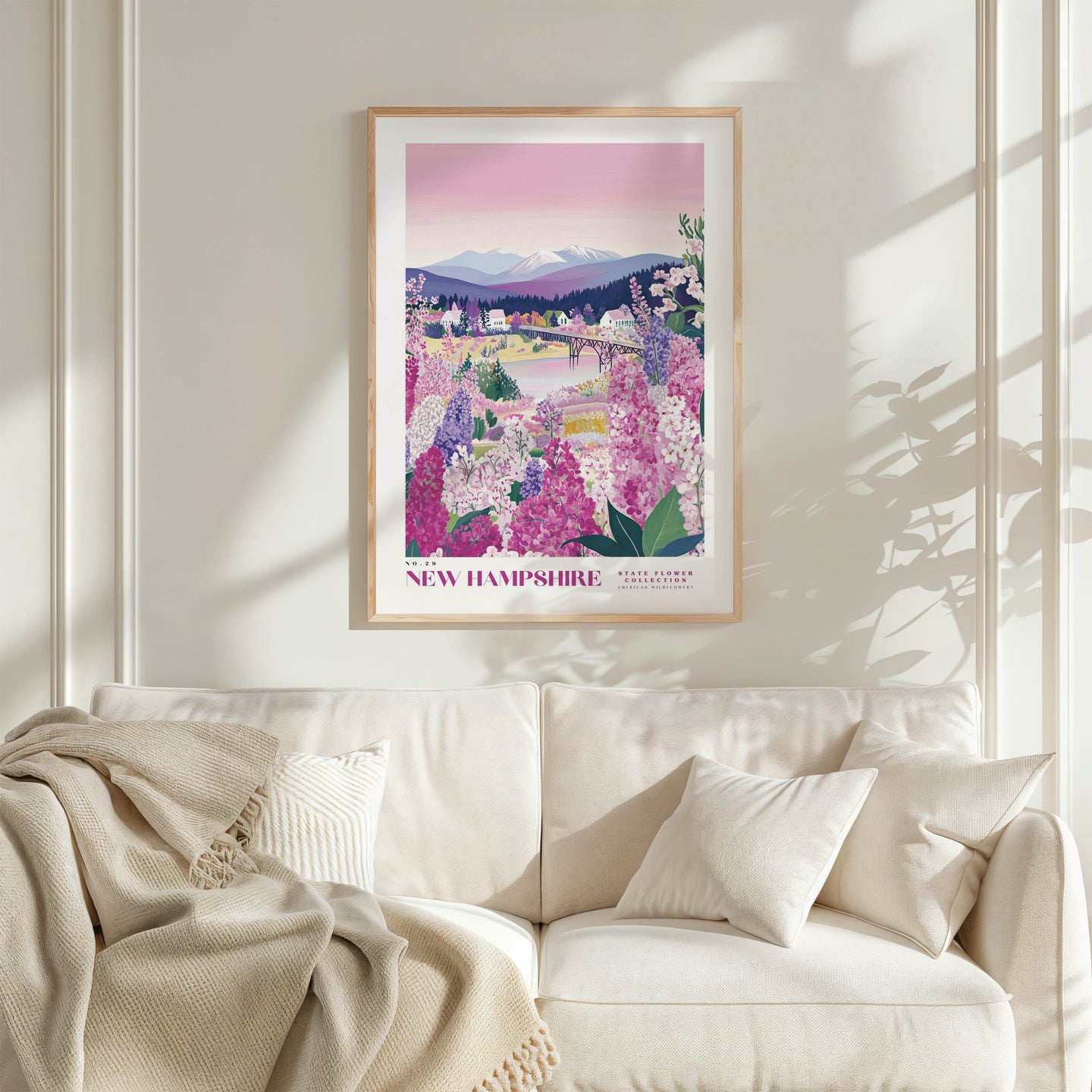 a framed poster of a scenic landscape with mountains, flowers, and a river, hanging on a wall above a white couch with a blanket draped over it.
