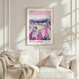 a framed poster of a scenic landscape with mountains, flowers, and a river, hanging on a wall above a white couch with a blanket draped over it.