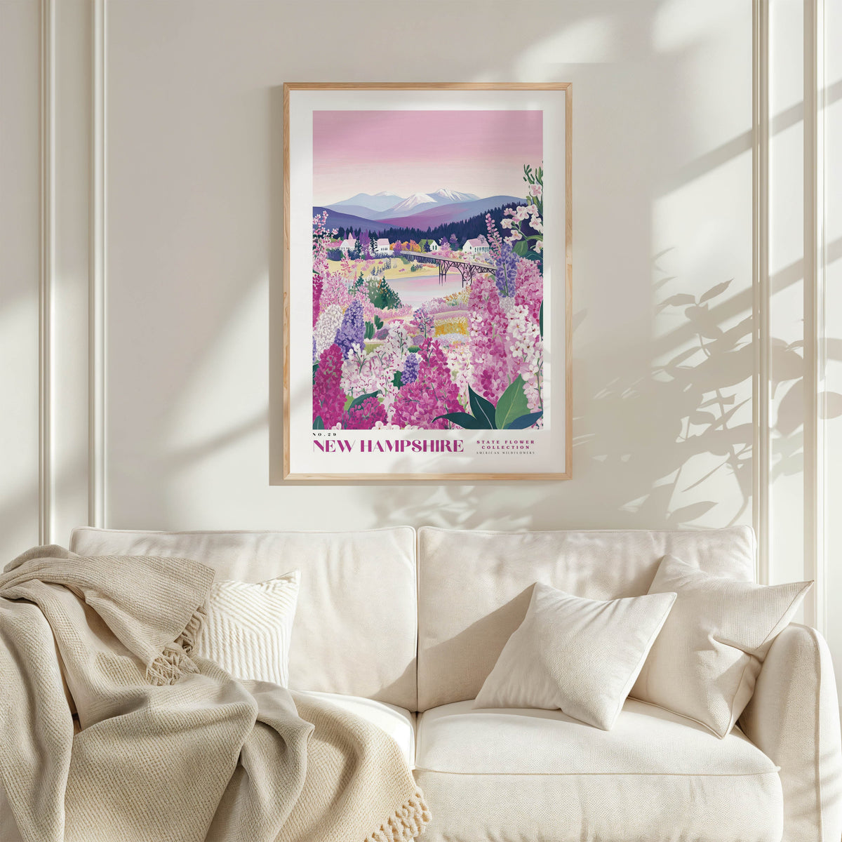 a framed poster of a scenic landscape with mountains, flowers, and a river, hanging on a wall above a white couch with a blanket draped over it.