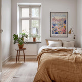 a cozy bedroom with a large bed, a wooden nightstand, and a window with a plant on the sill. The room has a neutral color scheme with a brown blanket and a colorful poster on the wall.