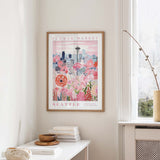 a framed poster of a cityscape with a flower market in the background, hanging on a wall in a room with a window and a small table with books and a vase.