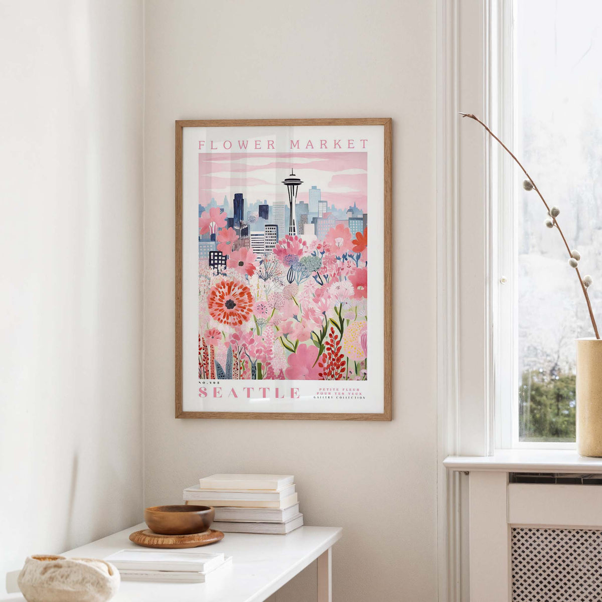 a framed poster of a cityscape with a flower market in the background, hanging on a wall in a room with a window and a small table with books and a vase.
