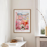 a framed poster of a colorful floral scene hanging on a wall, with a white table and a vase of flowers nearby.
