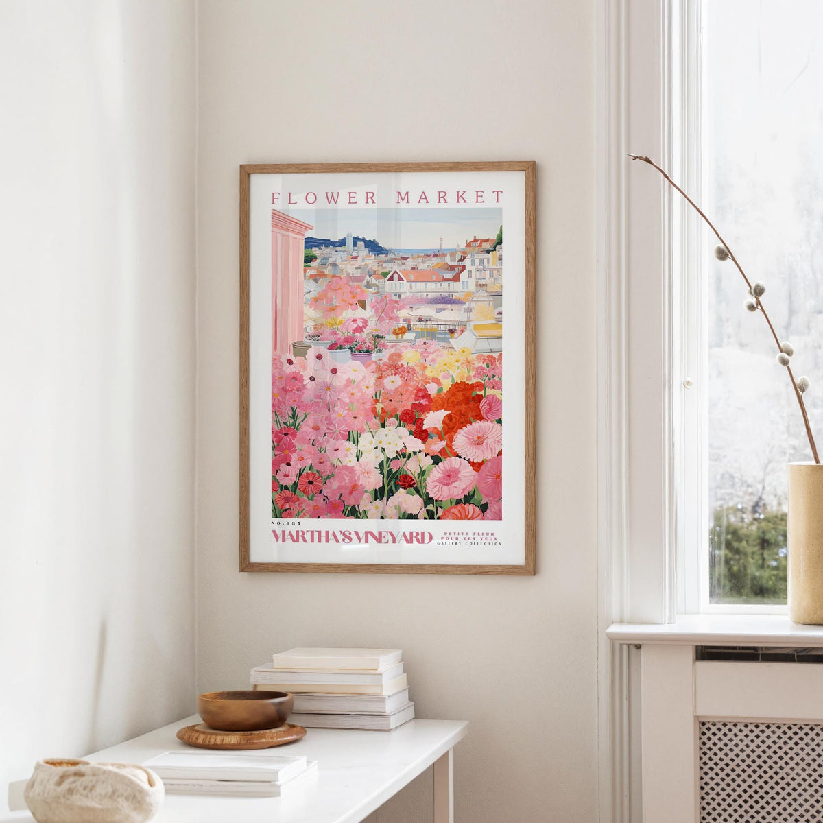 a framed poster of a colorful floral scene hanging on a wall, with a white table and a vase of flowers nearby.