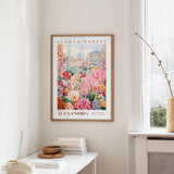 a framed poster of a colorful floral scene hanging on a wall, with a window and a small table with books and a vase nearby.