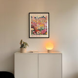 a white cabinet with a potted plant and a lamp on top, set against a wall with a framed poster of a cityscape above it.