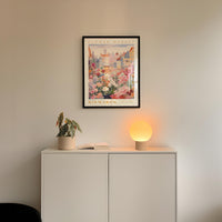 a white cabinet with a potted plant and a lamp on top, set against a wall with a framed poster of a cityscape above it.