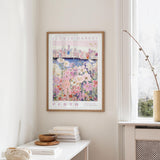 a framed poster of a cityscape with a flower market in the background, hanging on a wall in a room with a window and a small table with books and a bowl on it.