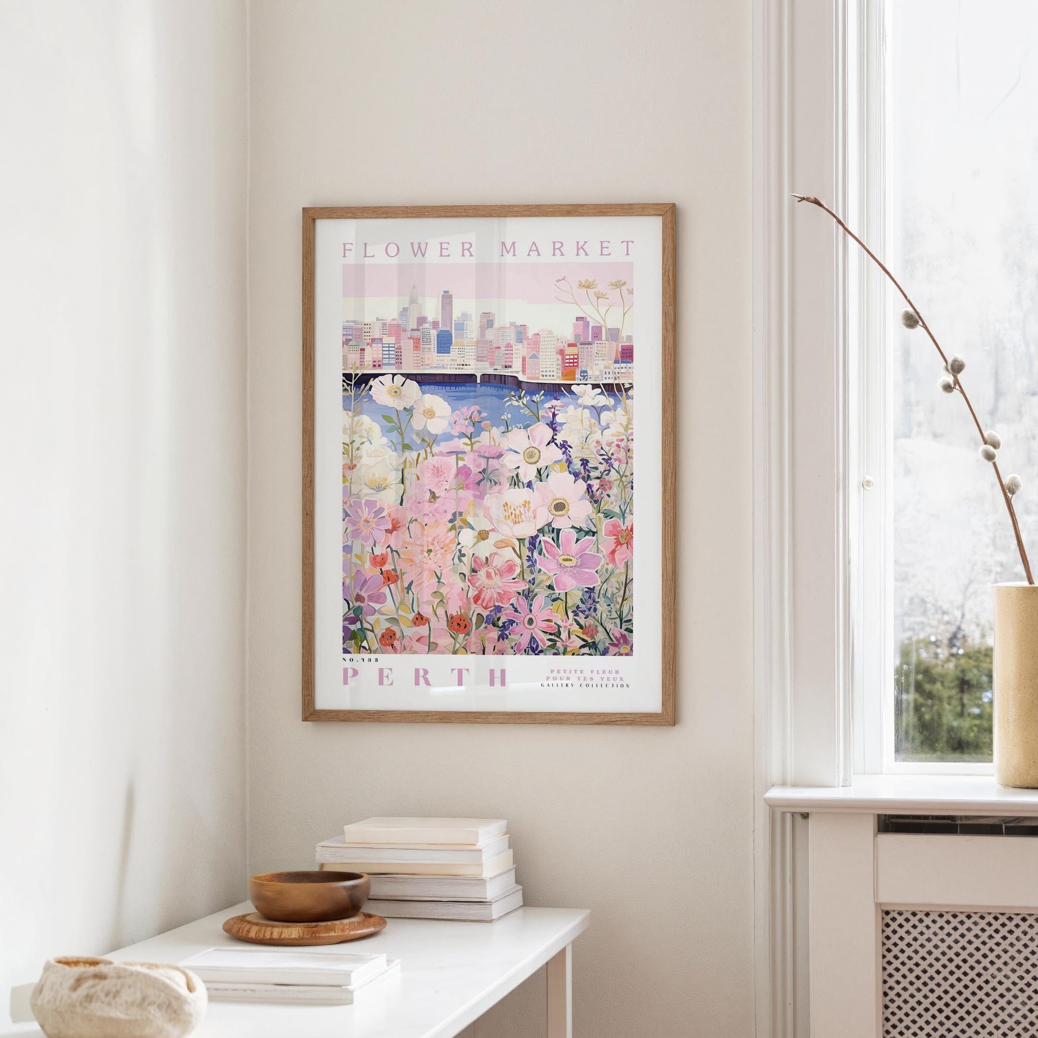 a framed poster of a cityscape with a flower market in the background, hanging on a wall in a room with a window and a small table with books and a bowl on it.