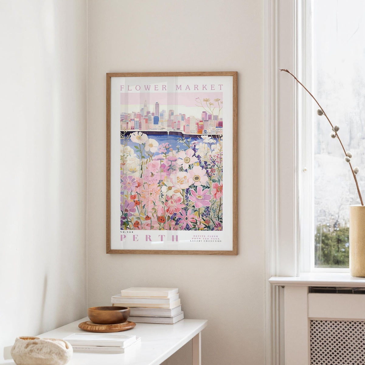 a framed poster of a cityscape with a flower market in the background, hanging on a wall in a room with a window and a small table with books and a bowl on it.