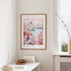 a framed poster of a cityscape with a floral design, hanging on a wall in a room with a window and a small table with books and a bowl on it.