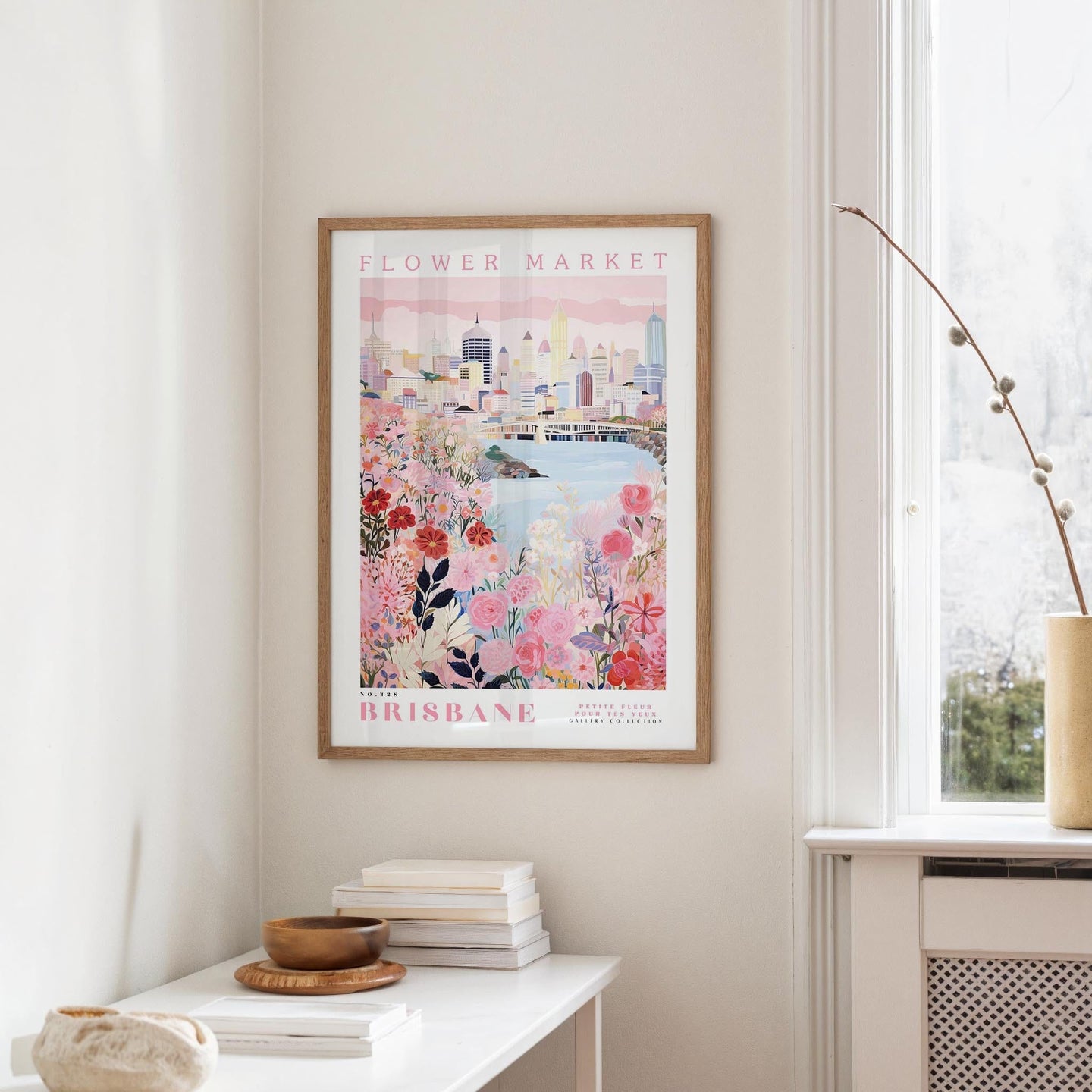 a framed poster of a cityscape with a floral design, hanging on a wall in a room with a window and a small table with books and a bowl on it.