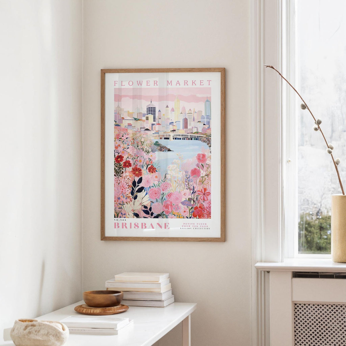 a framed poster of a cityscape with a floral design, hanging on a wall in a room with a window and a small table with books and a bowl on it.