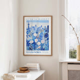 a framed poster of a cityscape with a flower market in the background, hanging on a wall in a room with a window and a small table with books and a bowl.