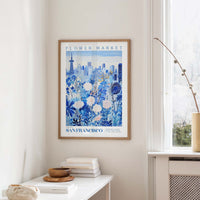 a framed poster of a cityscape with a flower market in the background, hanging on a wall in a room with a window and a small table with books and a bowl.