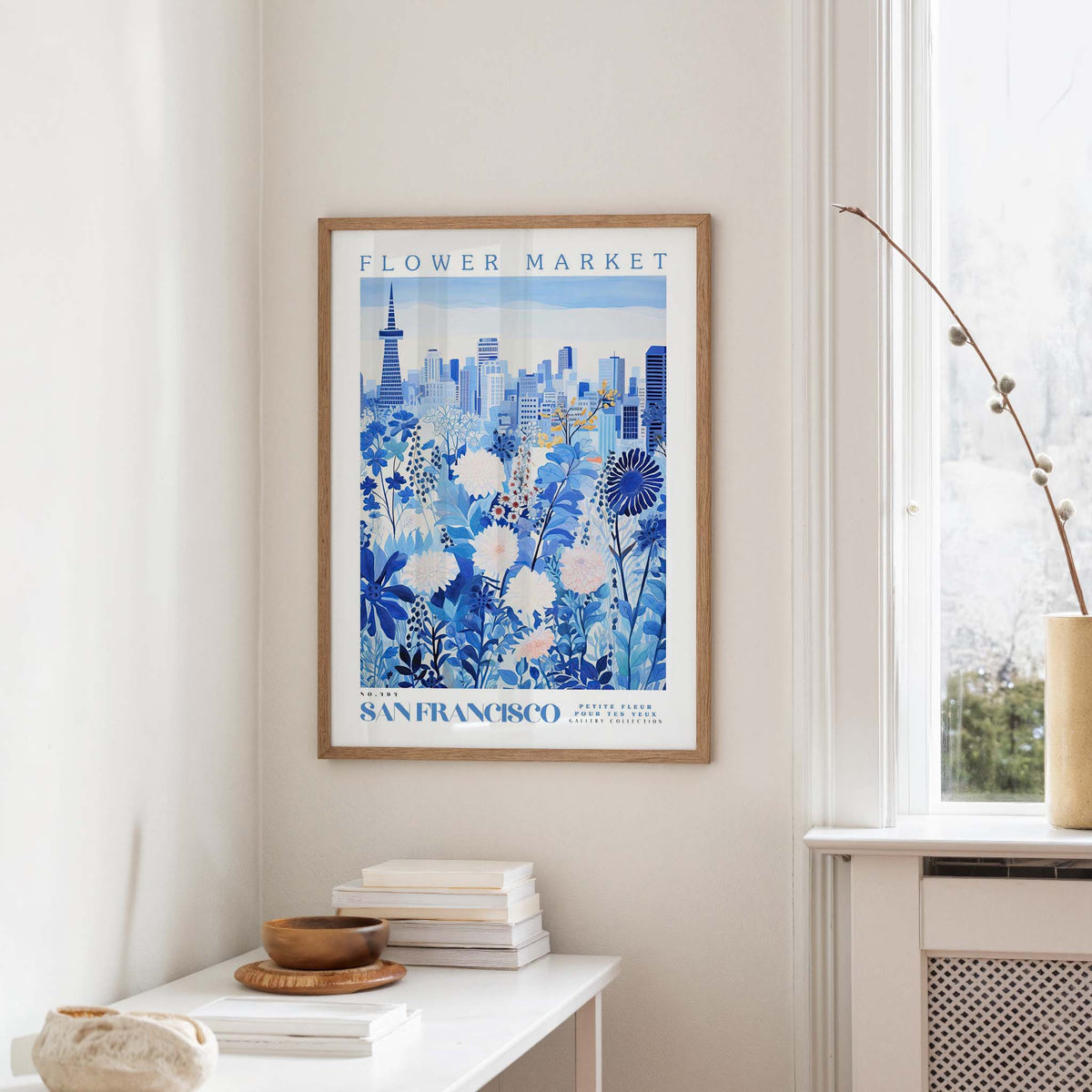 a framed poster of a cityscape with a flower market in the background, hanging on a wall in a room with a window and a small table with books and a bowl.