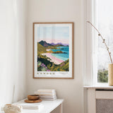 a framed poster of a scenic landscape with mountains, a beach, and a body of water, hanging on a wall in a room with a window and a small table with books and a vase.